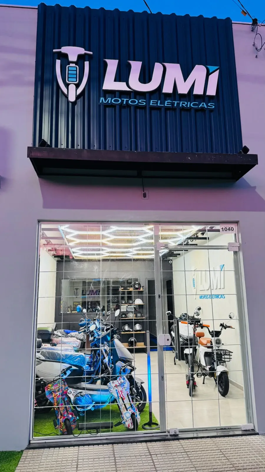 Fachada da loja LUMI Motos Elétricas iluminada ao entardecer, com o showroom visível pela vitrine